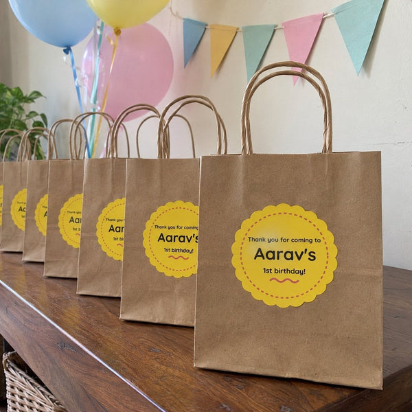 Kraft return-gift bags with yellow name stickers lined up on a table at a first-birthday party