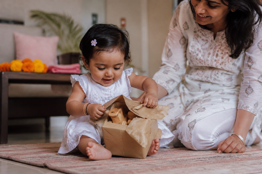 Indian baby girl about 12 months old sitting on a floor mat on her first birthday, pulling brown paper off a wrapped wooden toy
