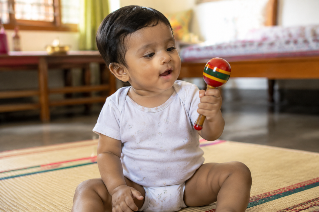 Indian baby around eight months old sitting on a floor mat shaking a glossy lacquered Channapatna style wooden rattle