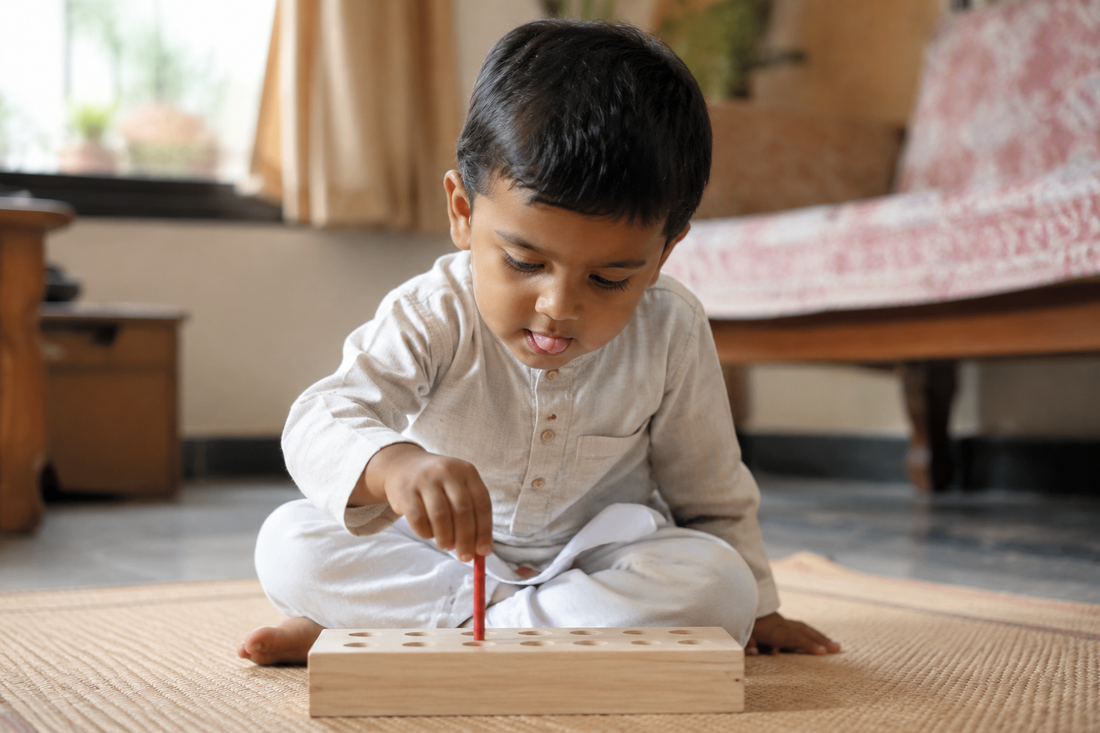 Indian toddler about two years old pinching a thin red wooden peg between thumb and finger, about to drop it into a hole in a pale wooden board