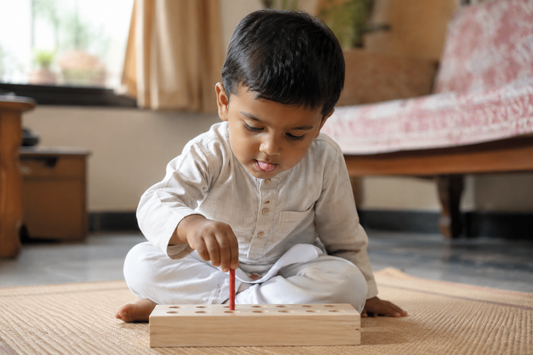 Indian toddler about two years old pinching a thin red wooden peg between thumb and finger, about to drop it into a hole in a pale wooden board