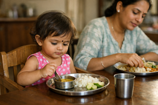Indian toddler about 2 years old sitting at the family dining table with a small steel thali, holding her own spoon, while her mother eats her own meal beside her