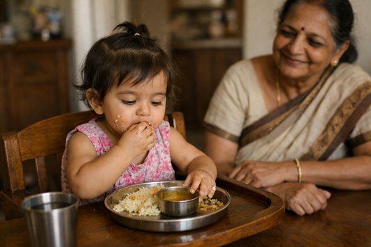 Indian toddler about 18 months old feeding herself with her fingers from a steel thali while her grandmother sits beside her without a spoon