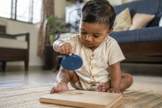 Indian baby about ten months old lifting a knobbed circular piece out of a pale wooden puzzle board on a floor mat