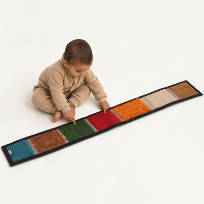 A toddler sitting on a white background, focused on pointing at the colorful and textured squares of the BrainyTots Sensory Texture Mat to develop fine motor skills.