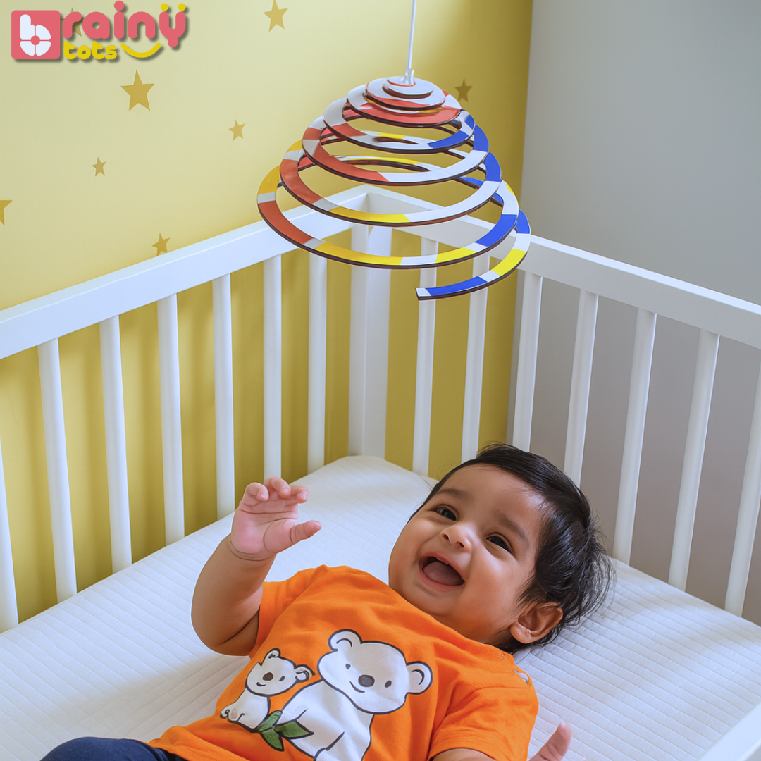 Happy baby reaching toward the BrainyTots Dual-Sided Spiral Hanging Mobile while visually tracking its gentle movement. This Montessori-inspired natural wooden baby mobile encourages visual development, focus, concentration, sensory exploration, and curiosity for newborns and babies aged 0–12 months.