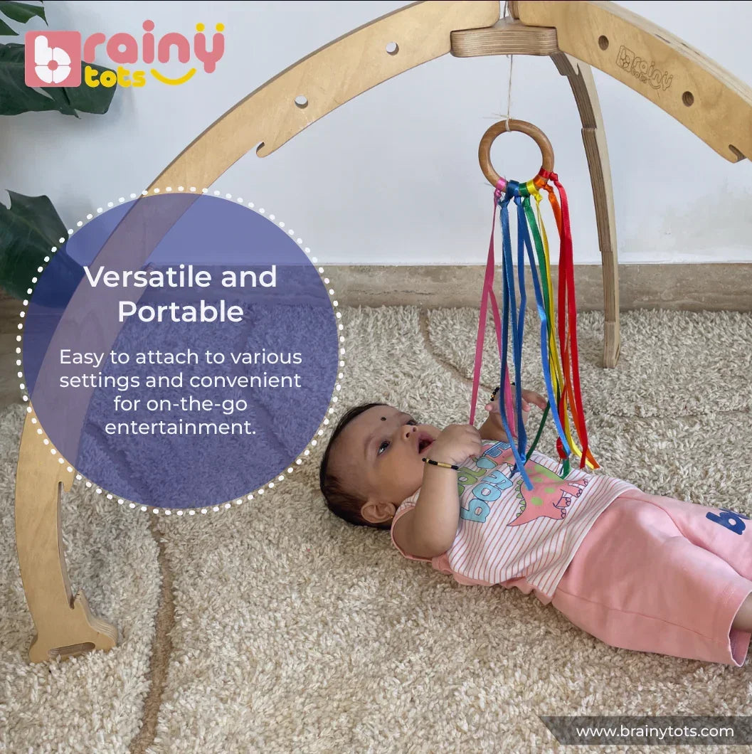 Baby playing with the BrainyTots Rainbow Ribbon Wooden Teether attached to a Montessori play gym, featuring a natural Neem wood ring and colorful ribbons that encourage sensory exploration, grasping, fine motor development, and portable play for babies aged 2 months and above.