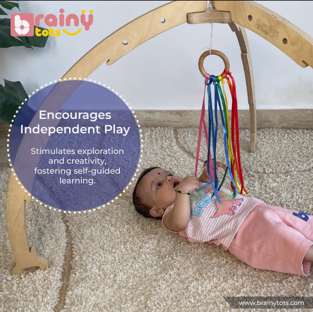 Baby independently exploring the BrainyTots Rainbow Ribbon Wooden Teether hanging from a Montessori play gym, using a natural Neem wood ring and colorful ribbons to encourage self-directed play, sensory exploration, grasping, hand-eye coordination, and fine motor development for babies aged 2 months and above