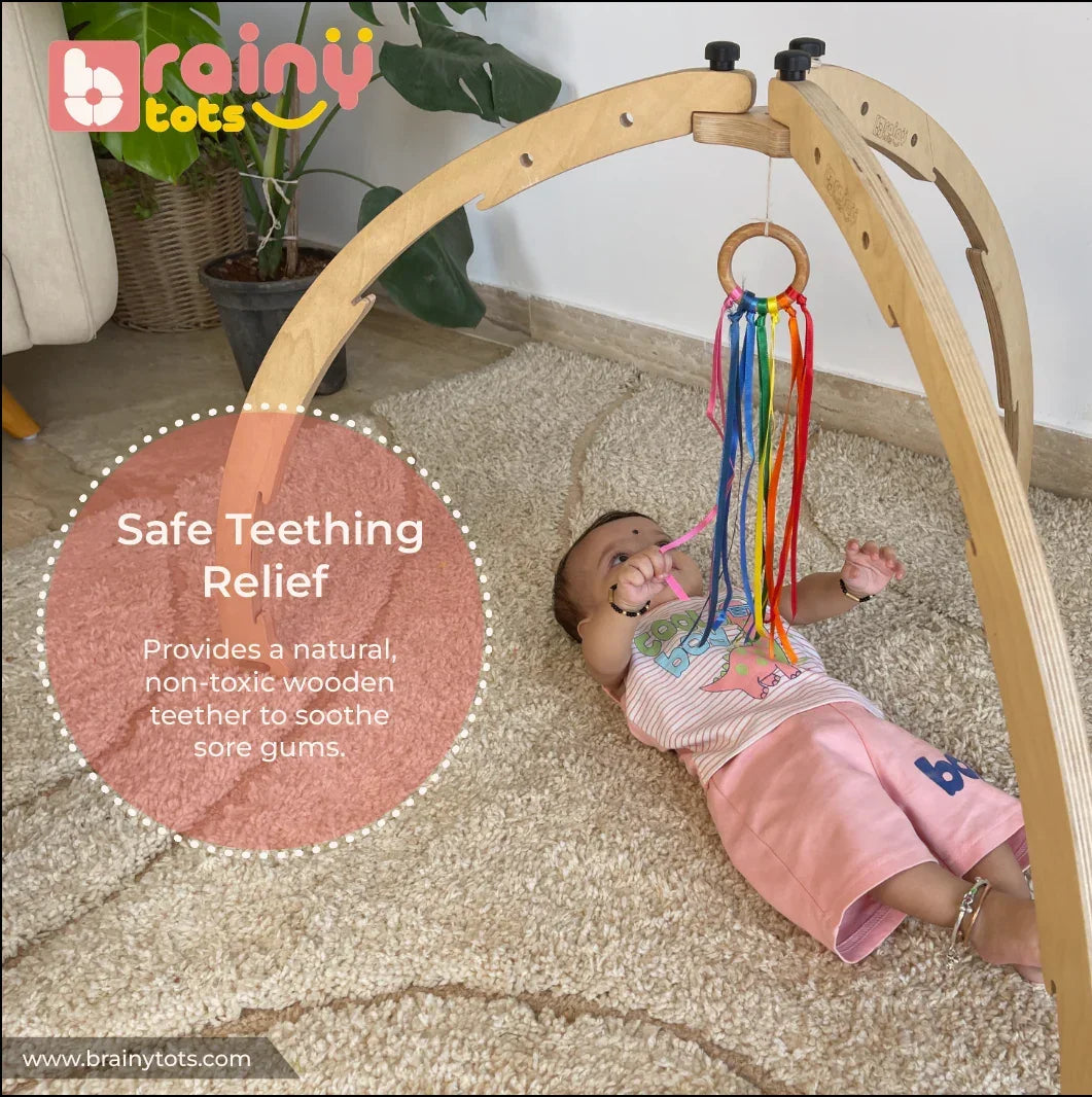 Baby playing with the BrainyTots Rainbow Ribbon Wooden Teether hanging from a Montessori play gym, providing natural teething relief while encouraging grasping, sensory exploration, hand-eye coordination, and fine motor development for babies aged 2 months and above.