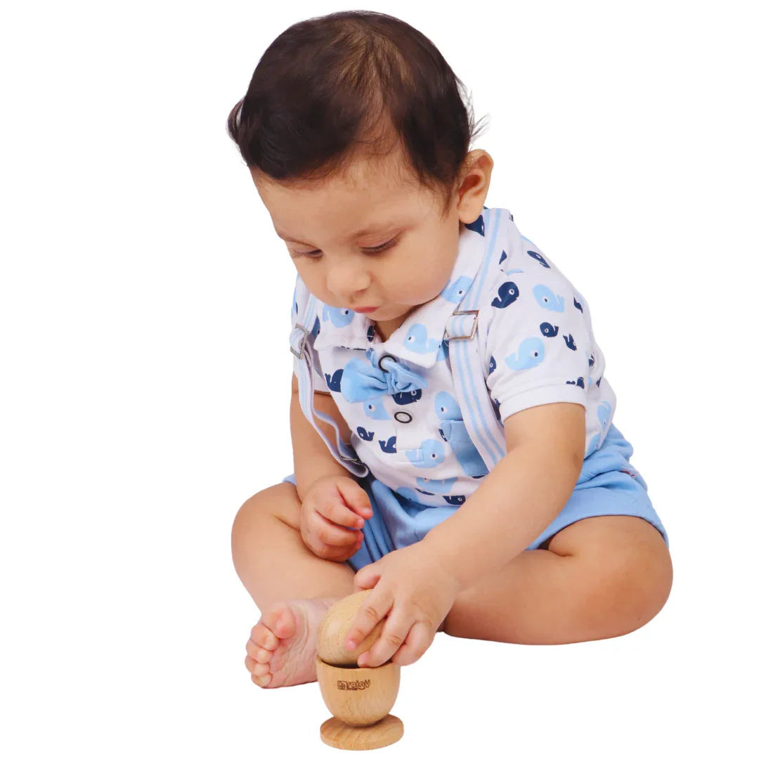 Baby sitting on a white background carefully placing a natural wooden egg into the cup piece of the Montessori puzzle, demonstrating balance, precision, and fine motor skill development for infants 6–15 months.
