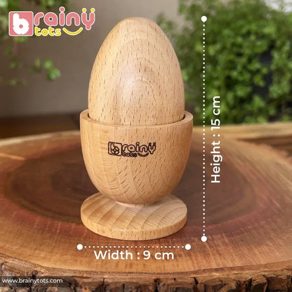 Assembled natural beech wood Montessori egg and cup puzzle with BrainyTots logo engraved on the cup, displayed on a rustic wood slice with green foliage background, showing dimensions of 15 cm height and 9 cm width for babies 6–15 months.
