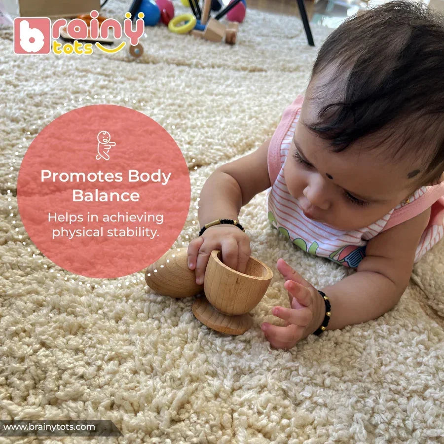 Baby doing tummy time on a soft rug while grasping a natural wooden Montessori egg and cup puzzle toy, with a coral overlay highlighting body balance and physical stability benefits for infants 6–15 months.
