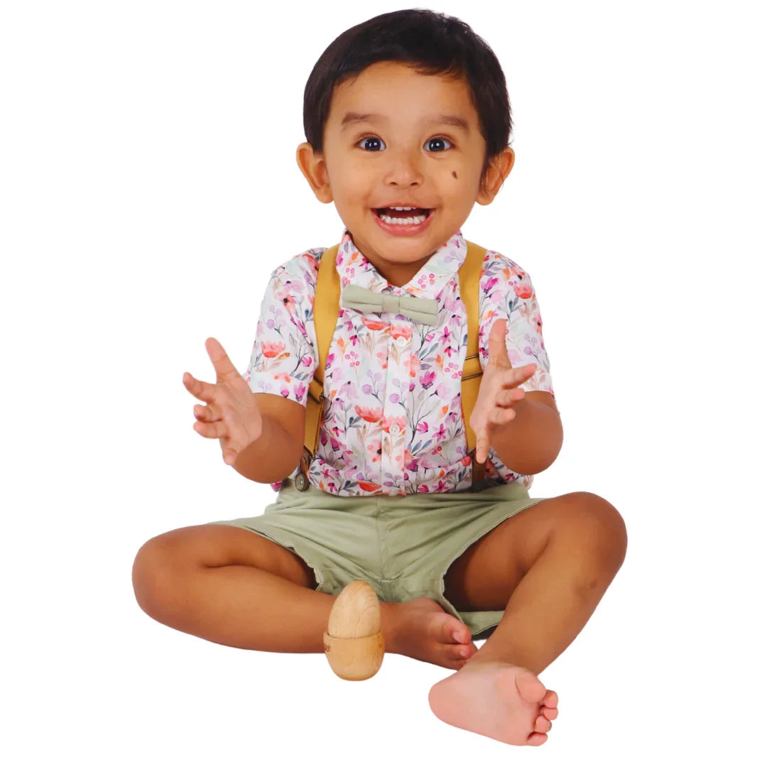 Happy toddler sitting cross-legged on a white background with hands raised in celebration, the natural wooden Montessori egg and cup puzzle assembled between their legs, capturing the joy of independent play success for babies 6–15 months.
