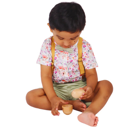 Toddler sitting cross-legged on a white background with head bowed in deep concentration, holding the natural wooden egg and reaching for the cup piece of the Montessori puzzle, demonstrating focused problem-solving and hand-eye coordination for babies 6–15 months.
