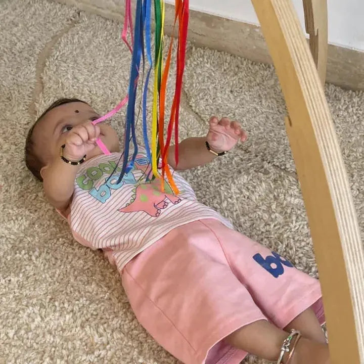 Baby lying under a play gym, reaching for the rainbow ribbon teether
